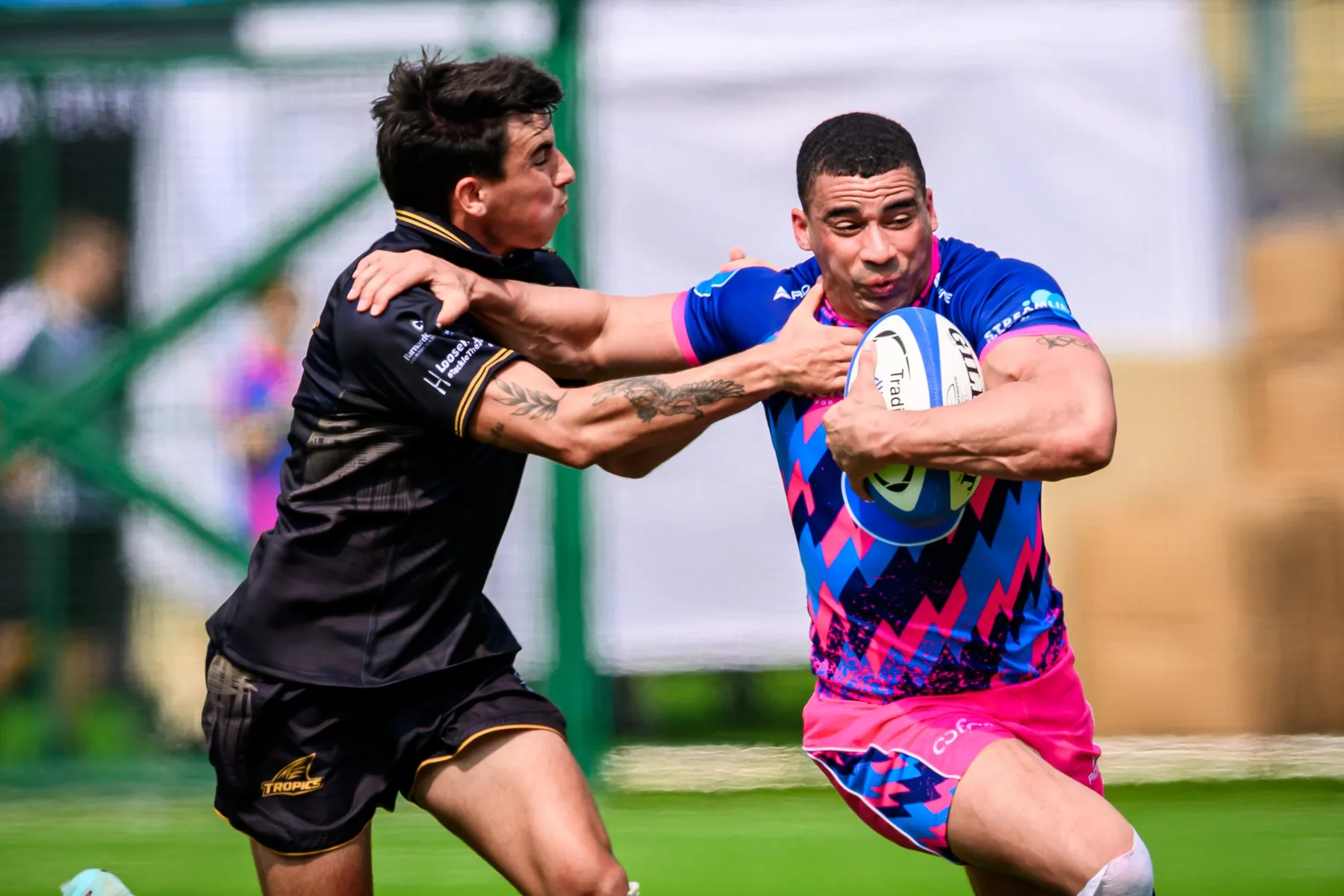 OSOB ball carrier in pink and blue fends off a Tropics defender in black with one outstretched arm