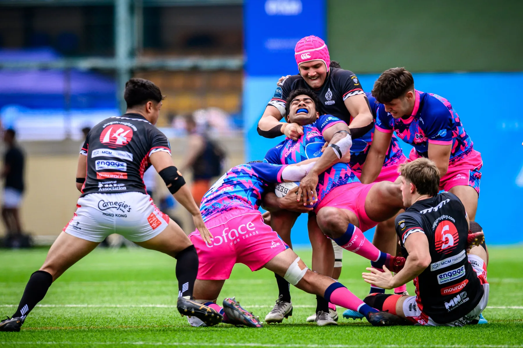 OSOB ball carrier is dragged down by black-shirted defenders in a crowded tackle, one opponent wearing a pink scrum cap