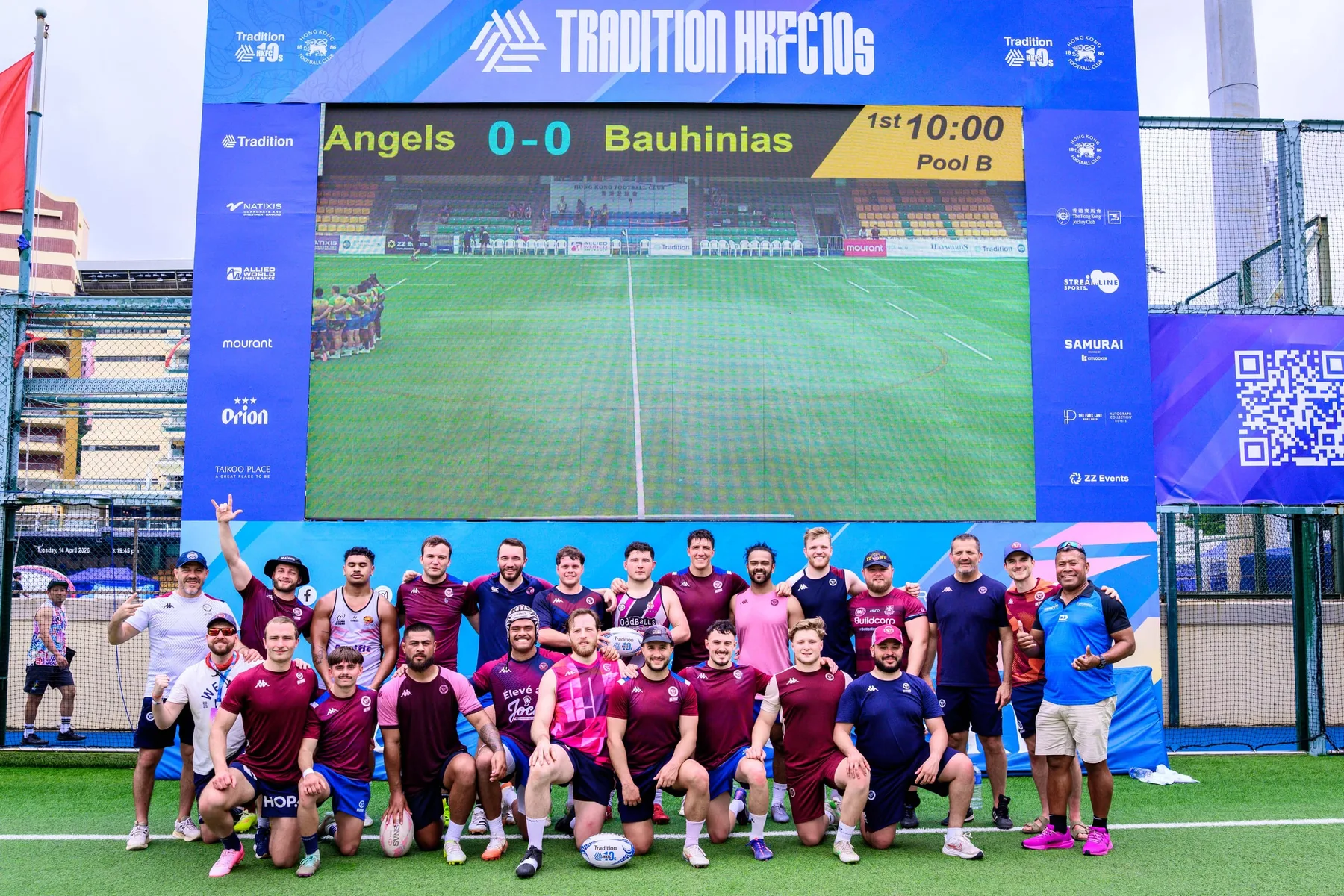 Squad in maroon shirts and a team-mate in pink pose before a big screen showing Angels against Bauhinias, 2026